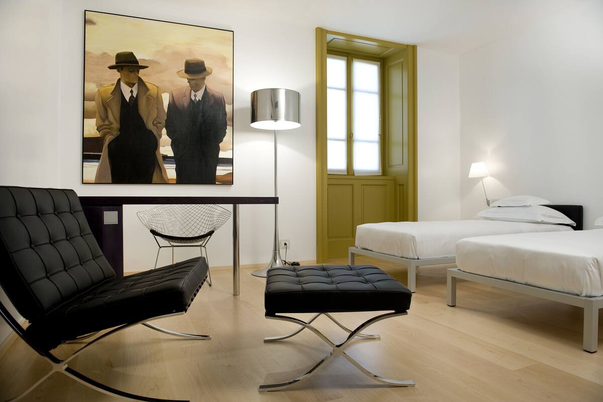 A modern hotel room with a black leather chair, a small table, two beds, a painting, and a floor lamp.