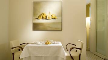 Un comedor con una mesa preparada para dos personas, un cuadro de peras en la pared y vistas al exterior a través de un ventanal.