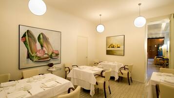 A dining area with tables set for four, white tablecloths, and chairs. There are paintings on the walls, and the room has a tiled floor.