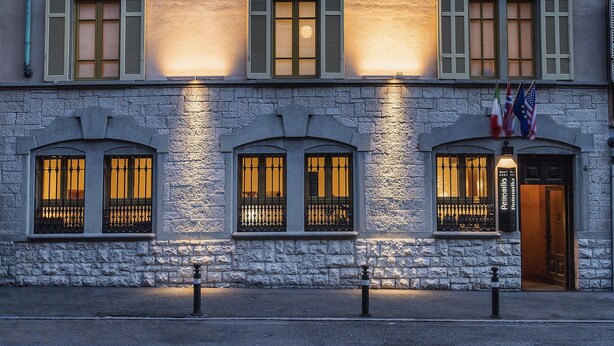 A building with the name "ALBERGO" illuminated on its facade, featuring multiple windows with shutters and a stone base.