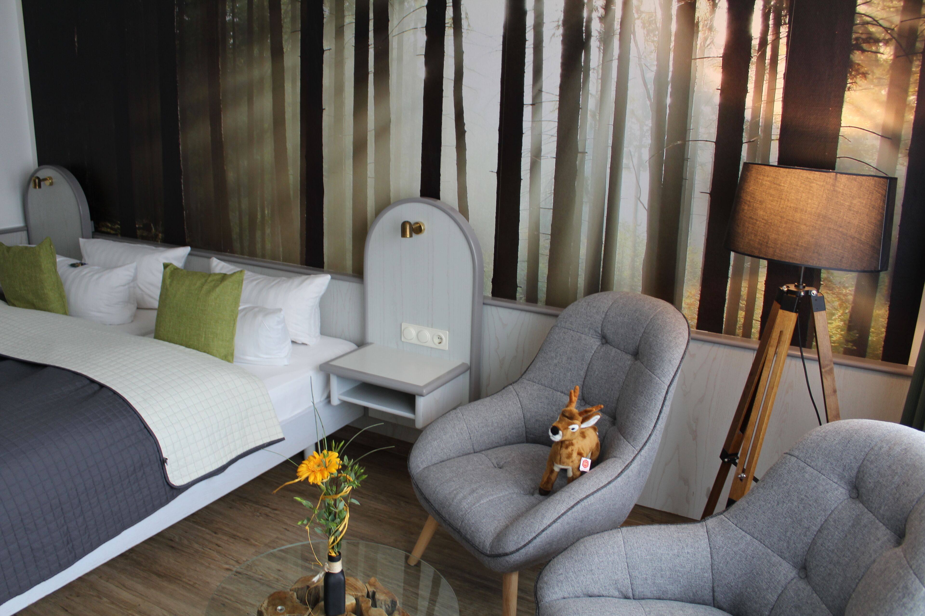 A hotel room with a bed, a gray armchair, a small table with a vase of flowers, and a painting of trees on the wall.