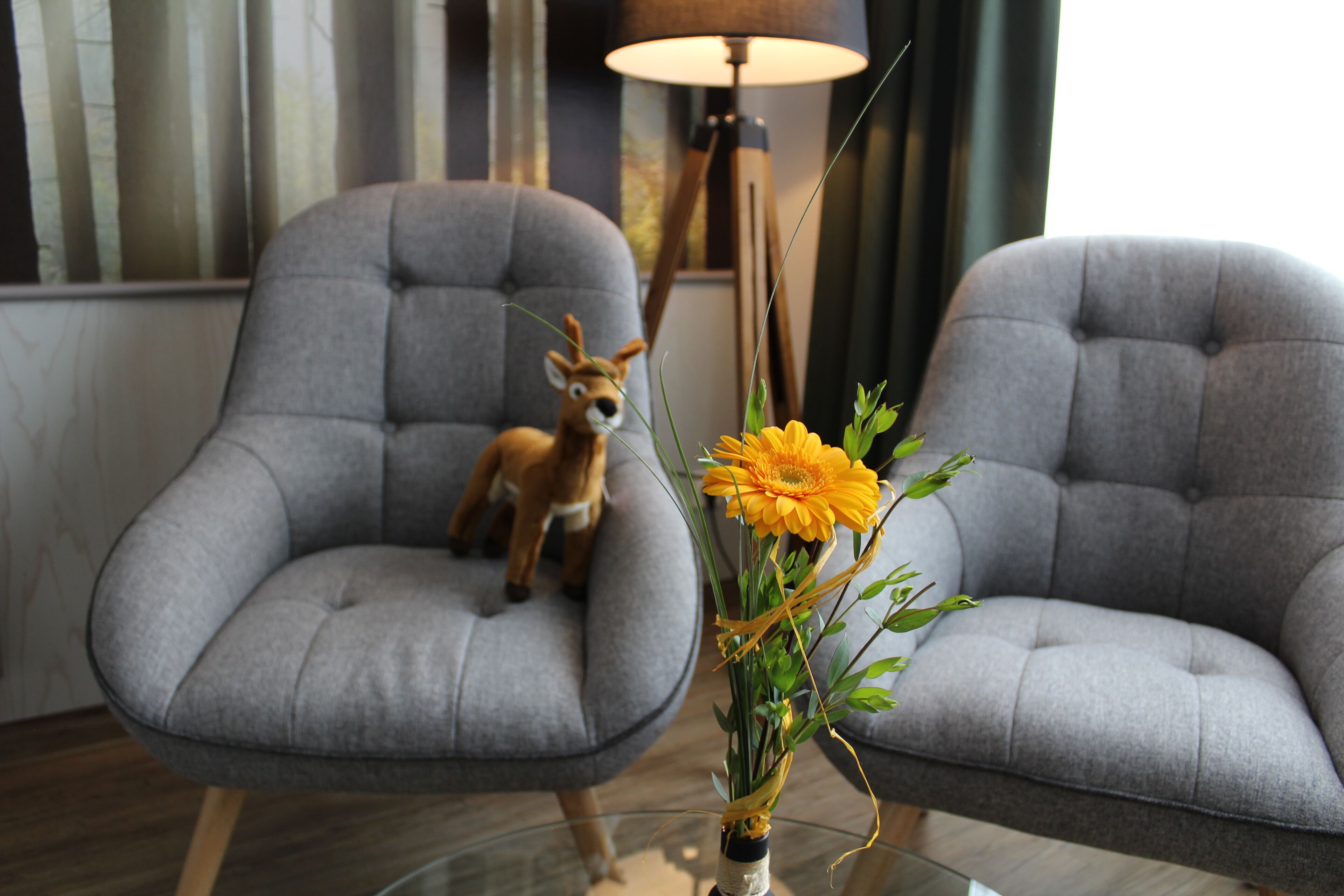 Two grey armchairs, a small deer figurine, and a vase with yellow flowers on a glass table.