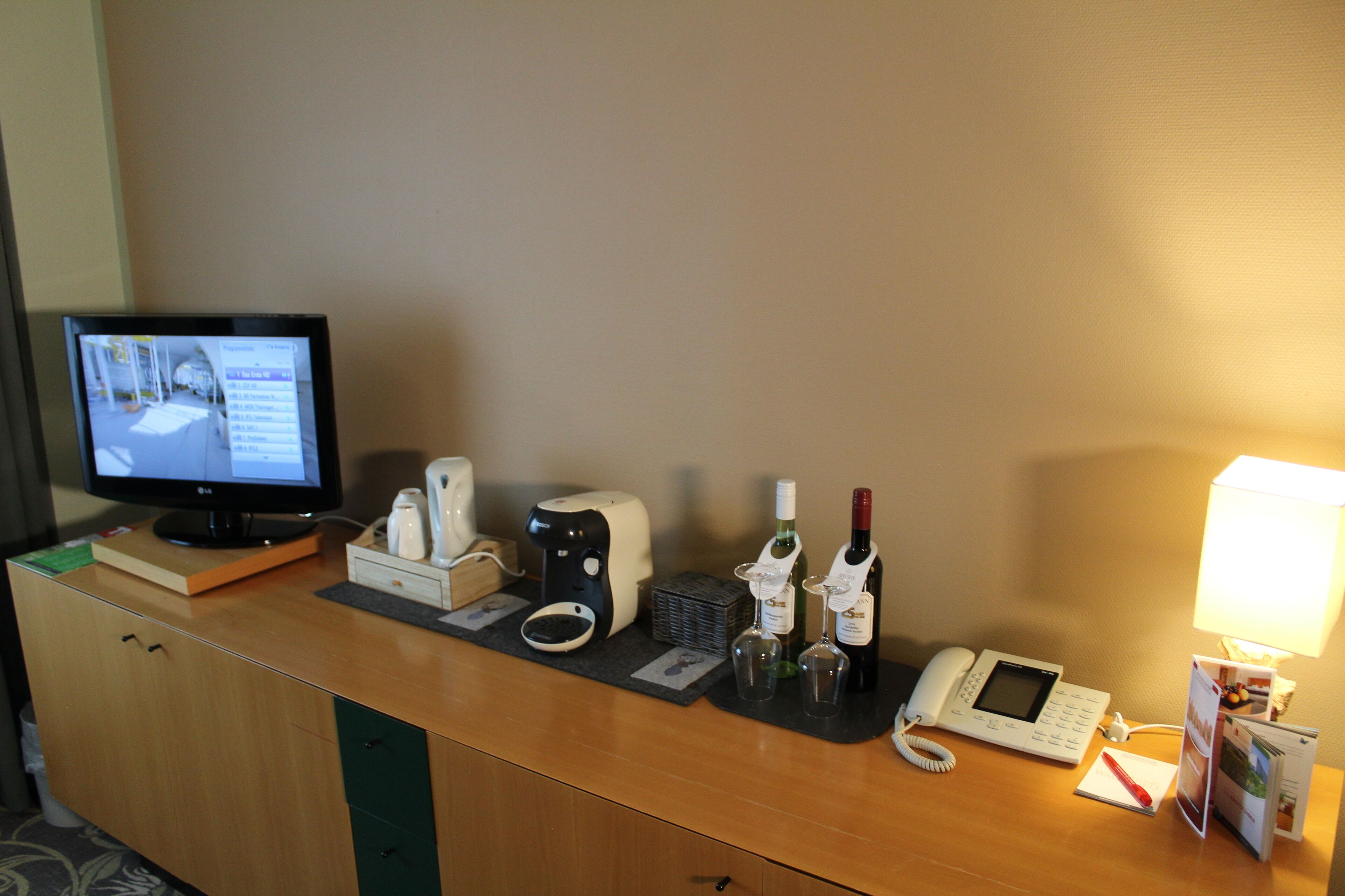 Mostrador de recepción de hotel con monitor de computadora, teléfono, botellas de vino y cafetera.