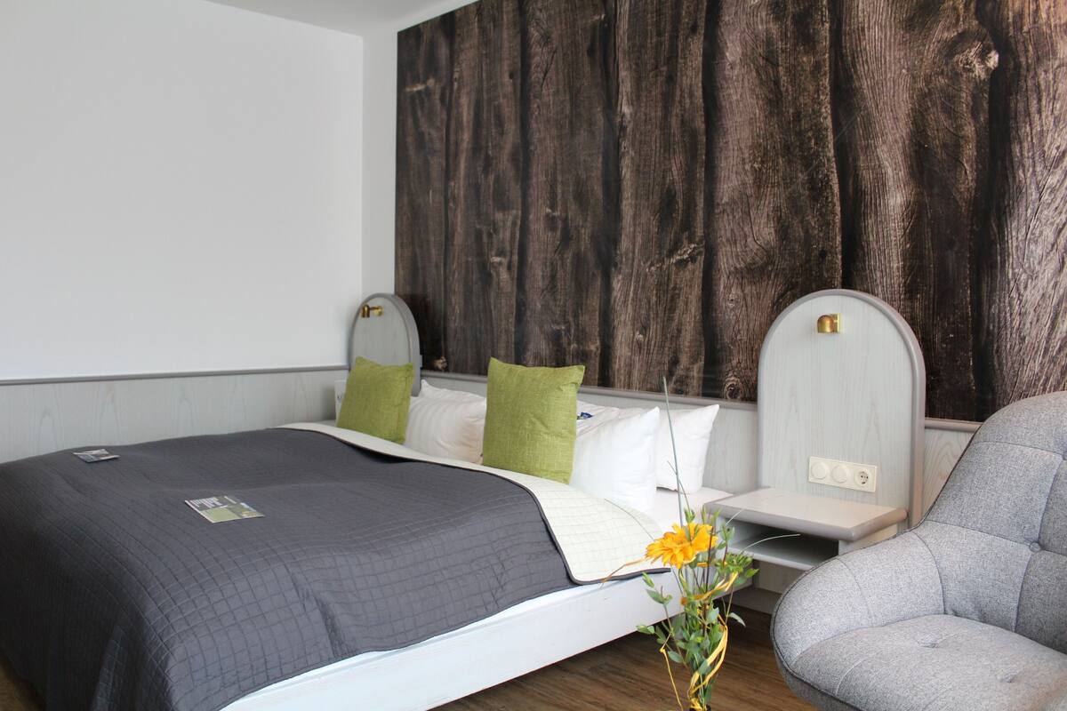 A modern hotel room with a large bed, a grey armchair, and a wooden accent wall.