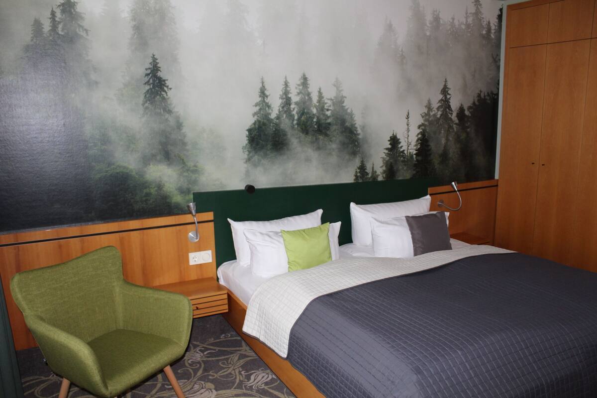 A hotel room with a bed, a green armchair, and a forest mural on the wall.
