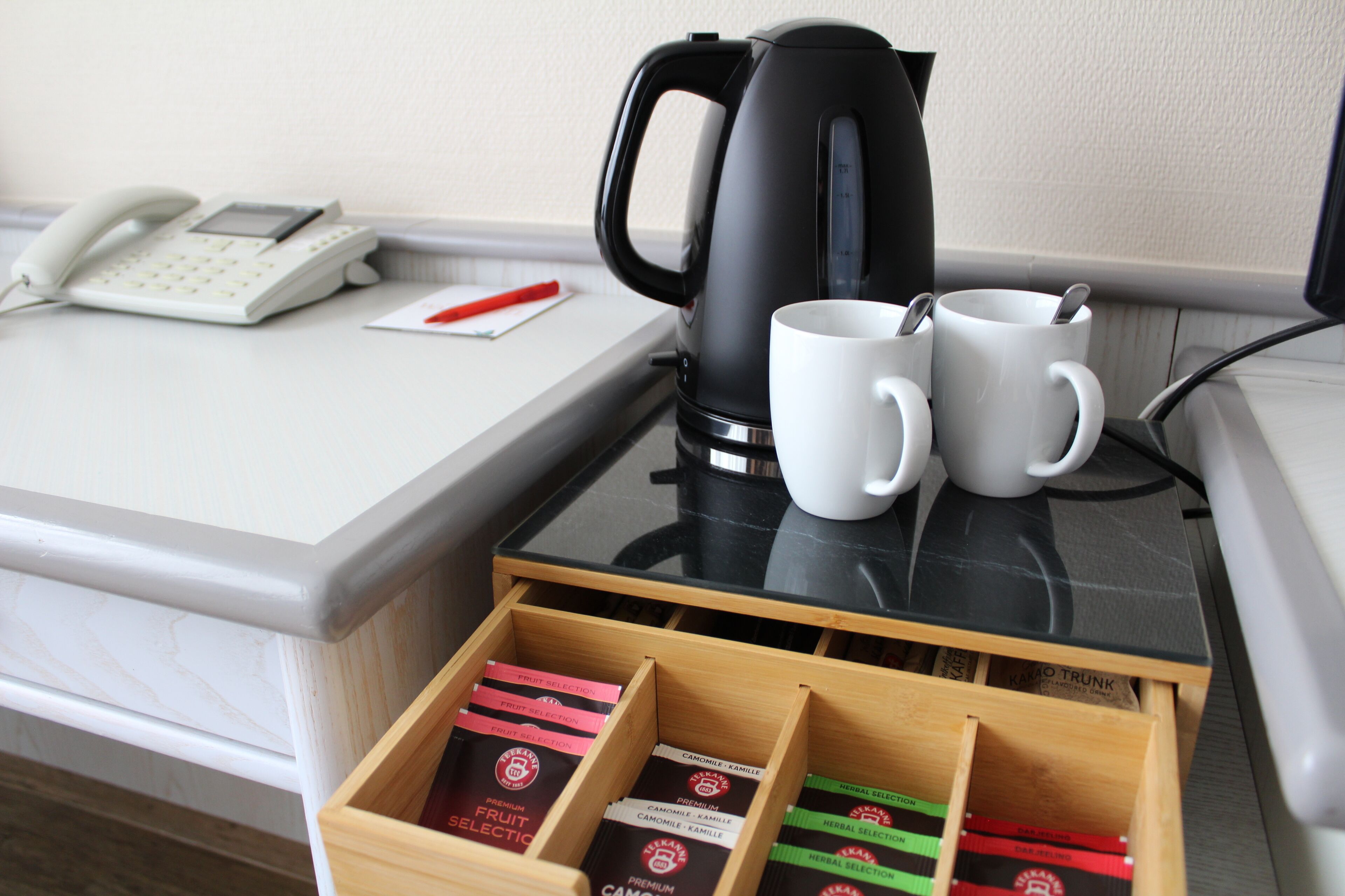 A hotel room with a desk, a telephone, a coffee maker, two mugs, and a tray of herbal tea boxes.