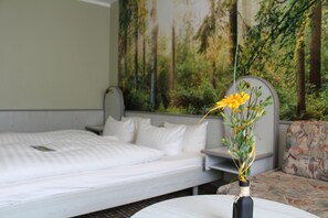 A hotel room with a bed, a white round table with a vase of flowers, and a mural of a forest on the wall.