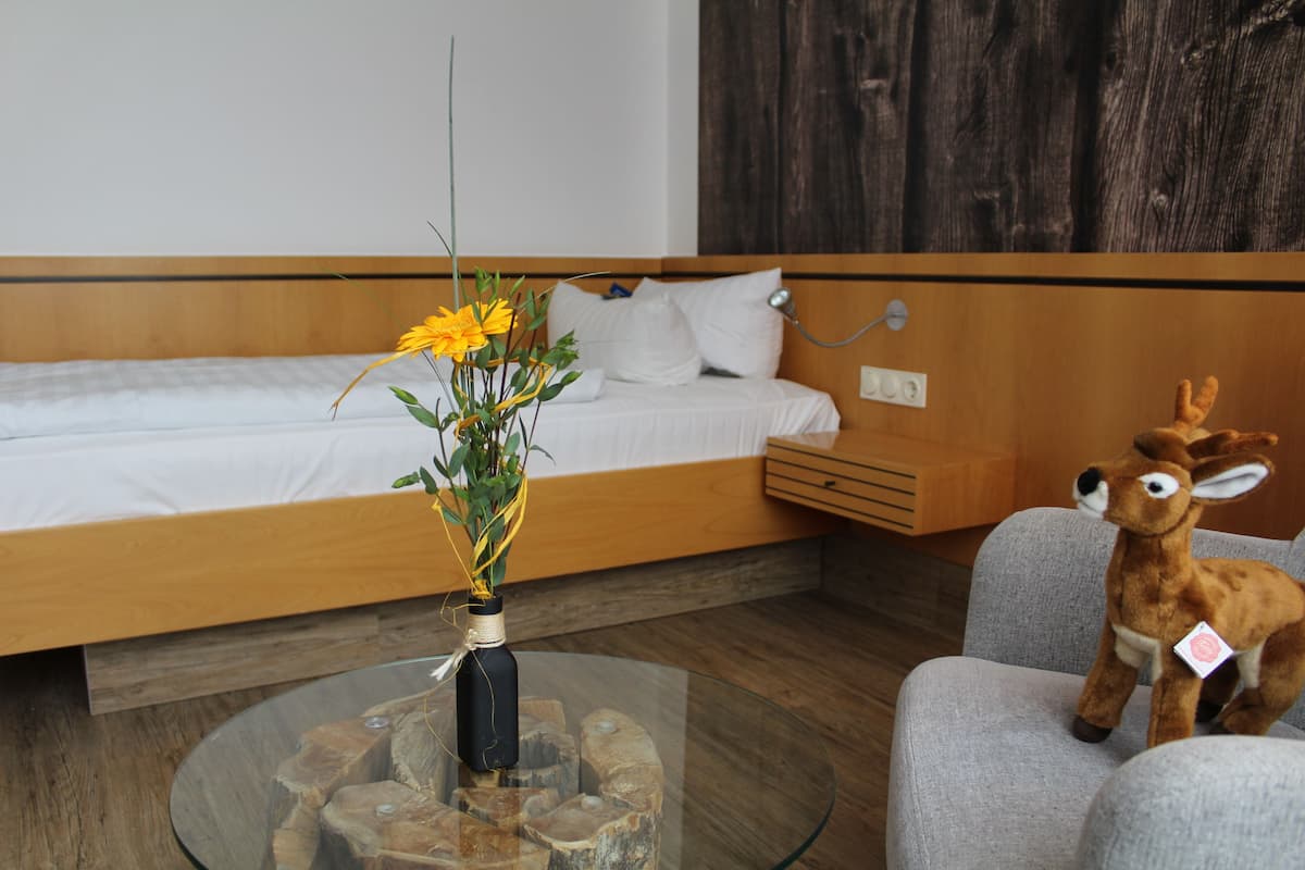 A modern hotel room with a wooden headboard, a glass coffee table with a vase of flowers, a plush deer toy, and a grey armchair.