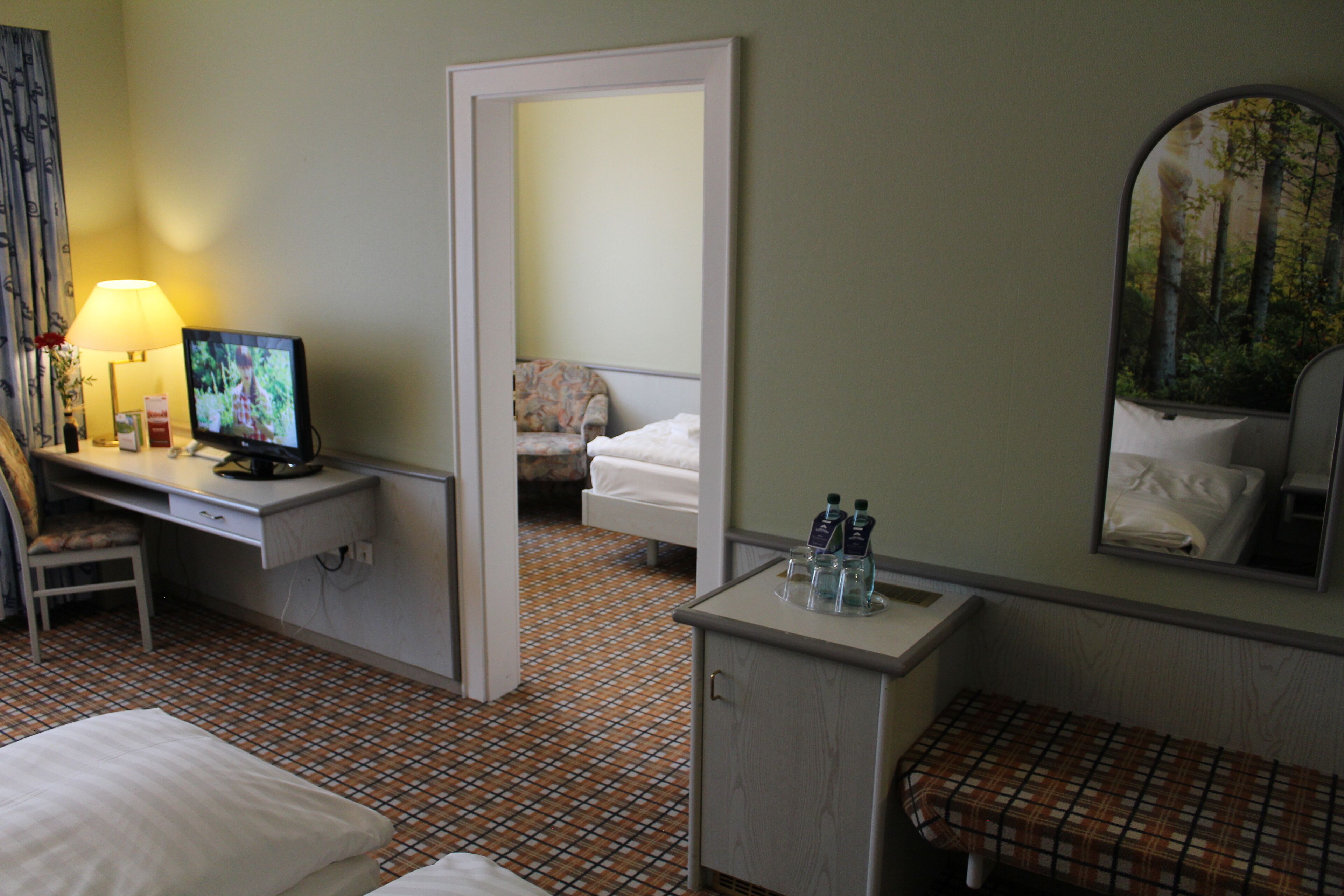 Et hotelværelse med en seng, et skrivebord med en computer, en stol, et lille bord med toiletartikler og et spejl der viser udsigt til et skovområde.