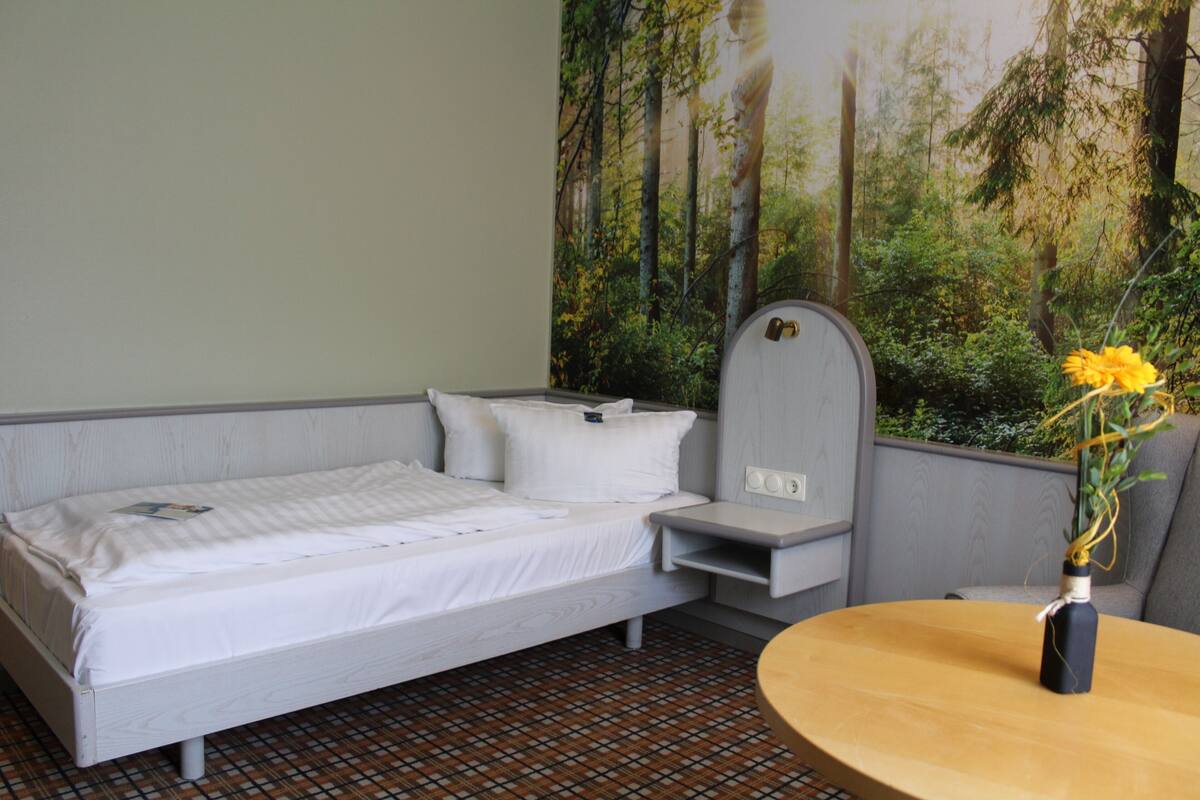 A bedroom with a bed, a small table with a vase, and a mural of a forest on the wall.