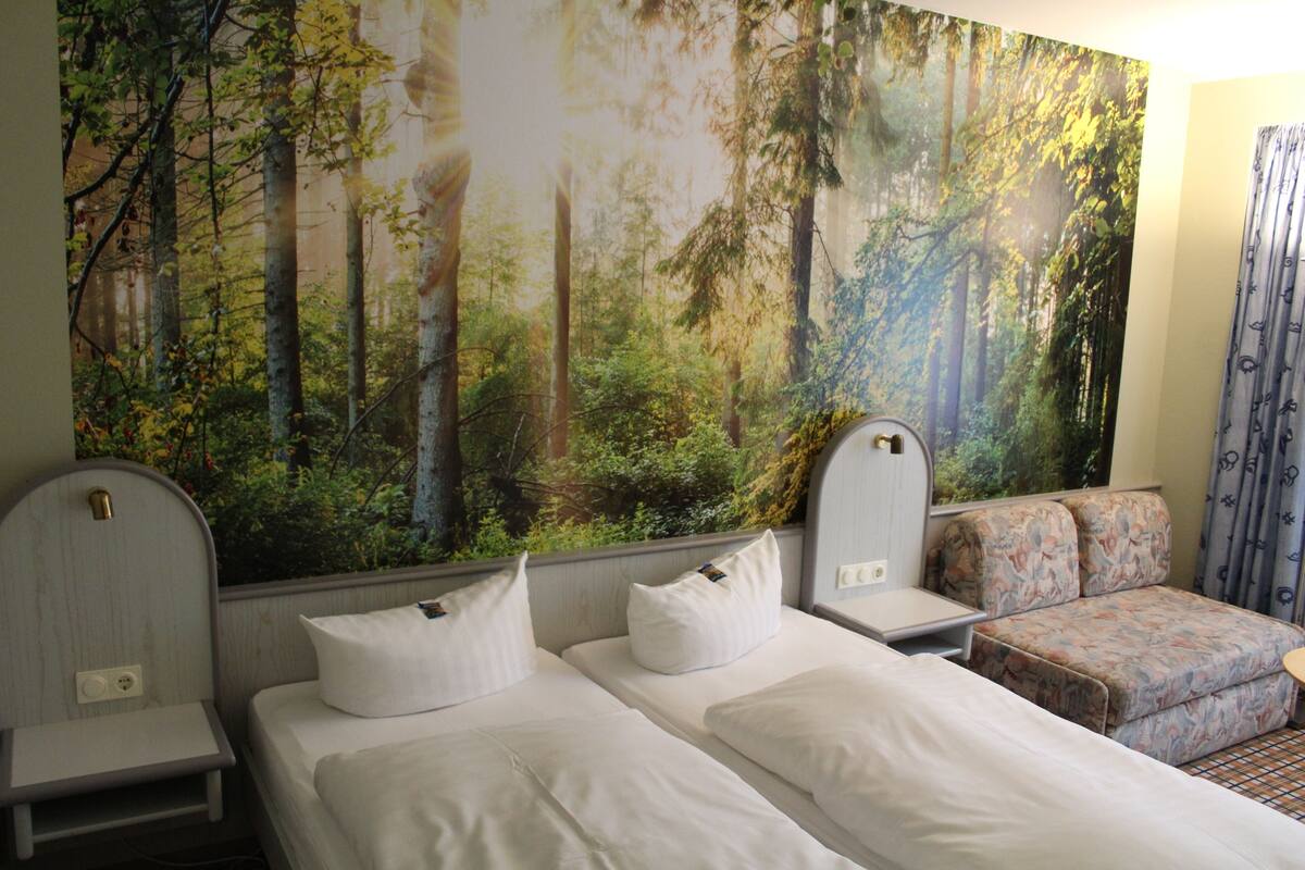 A hotel room with two beds, a sofa, and a large forest mural on the wall.