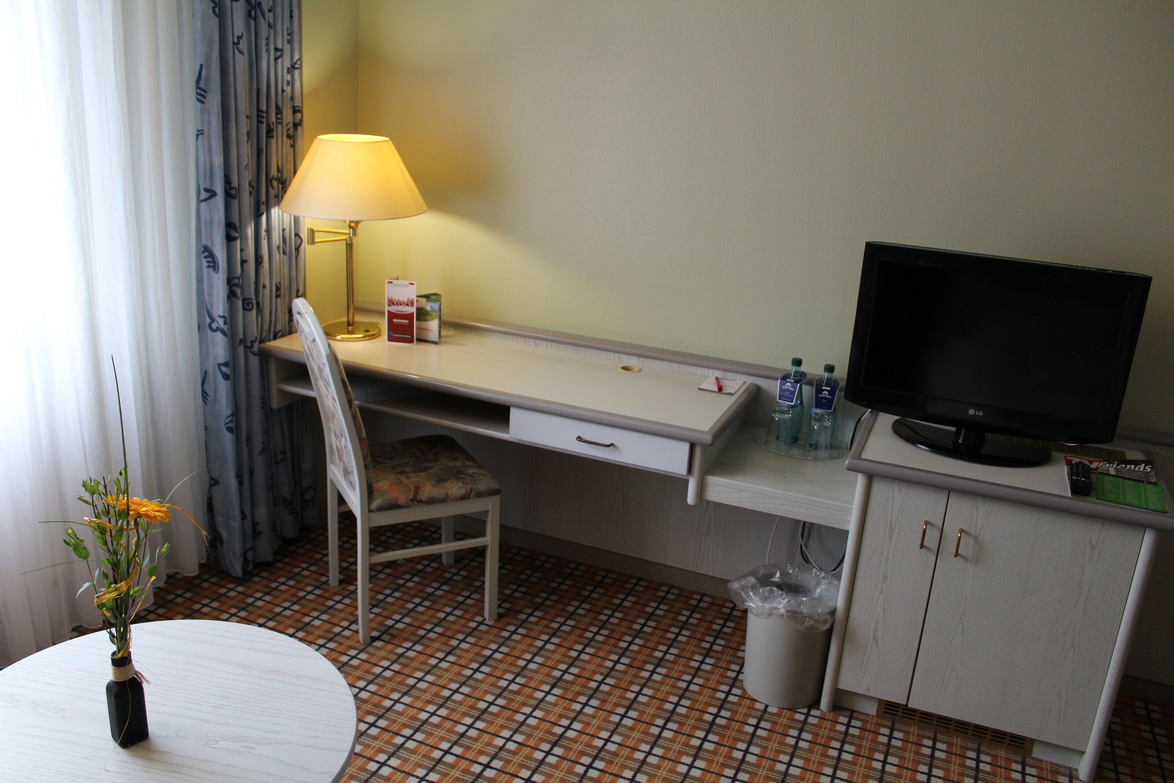 A hotel room with a desk, chair, TV, lamp, and a small table with a vase.