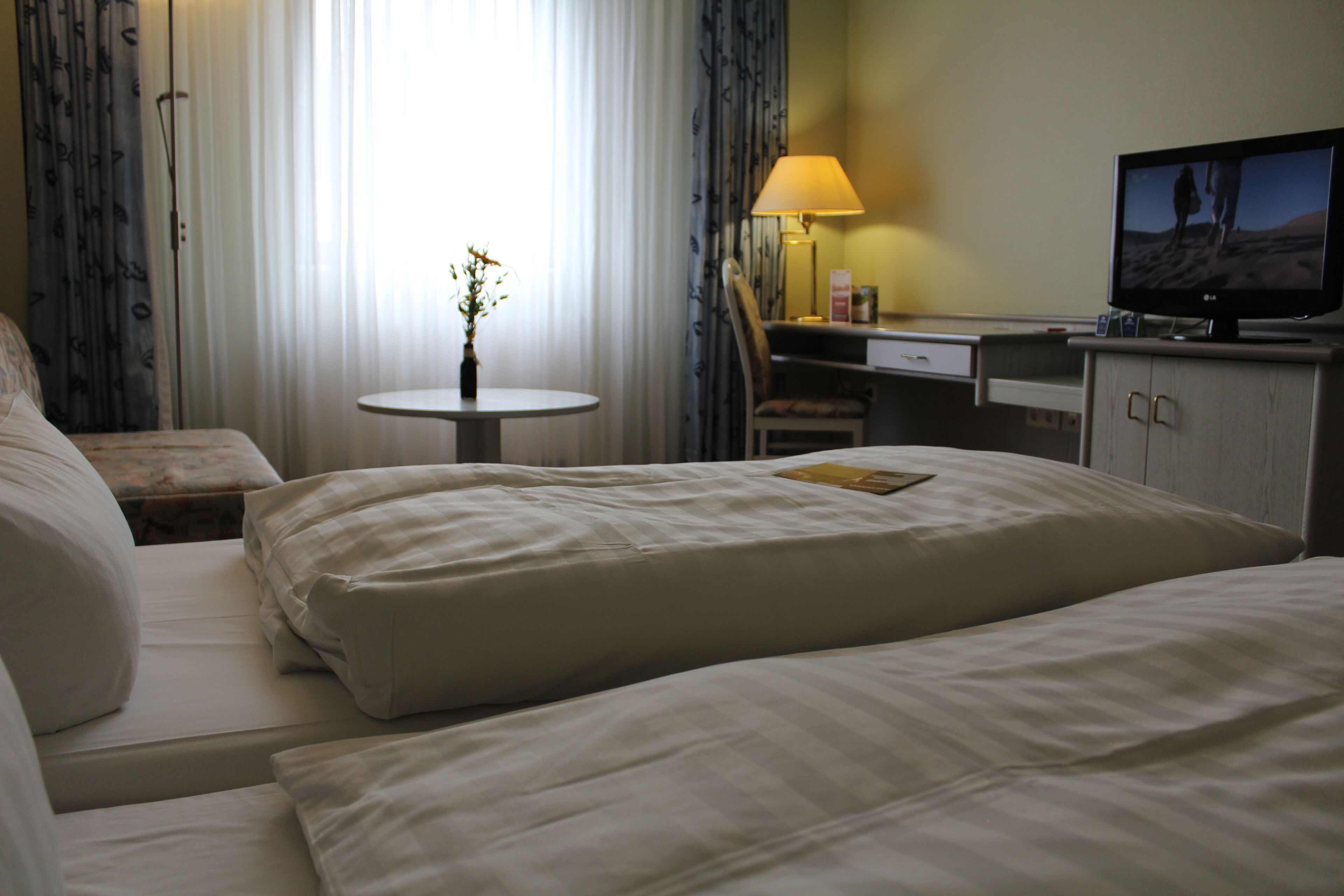 A hotel room with a bed, a desk with a lamp, a television, and a small table with a vase.