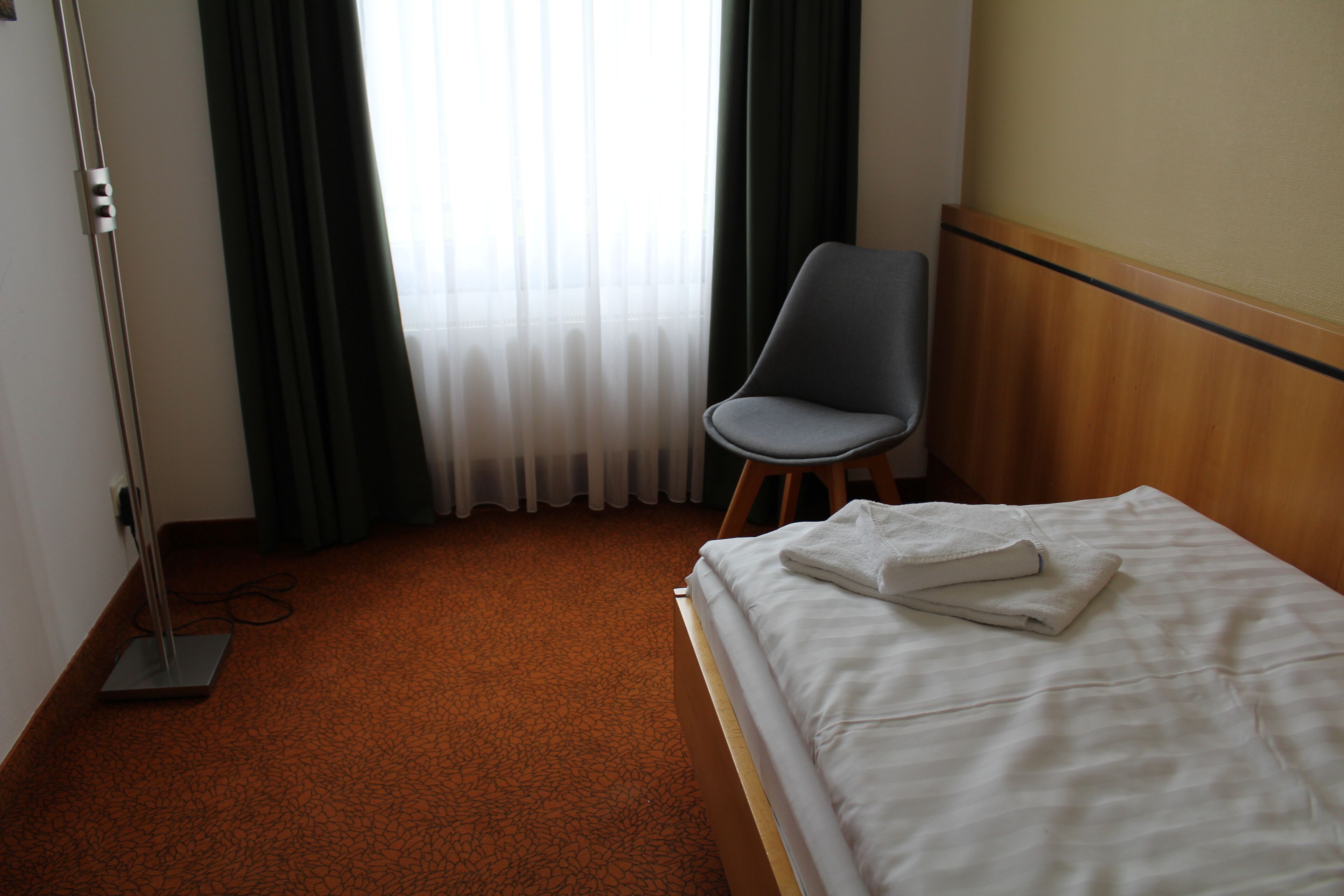 A hotel room with a bed, a chair, a floor lamp, and a window with curtains.