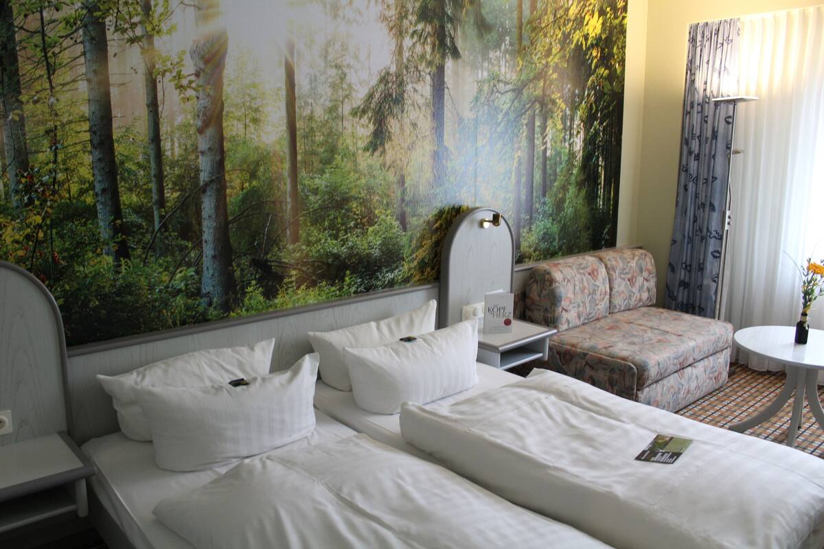 A hotel room with a large forest mural on the wall, a bed with white linens, a sofa, and a small round table with a vase of flowers.