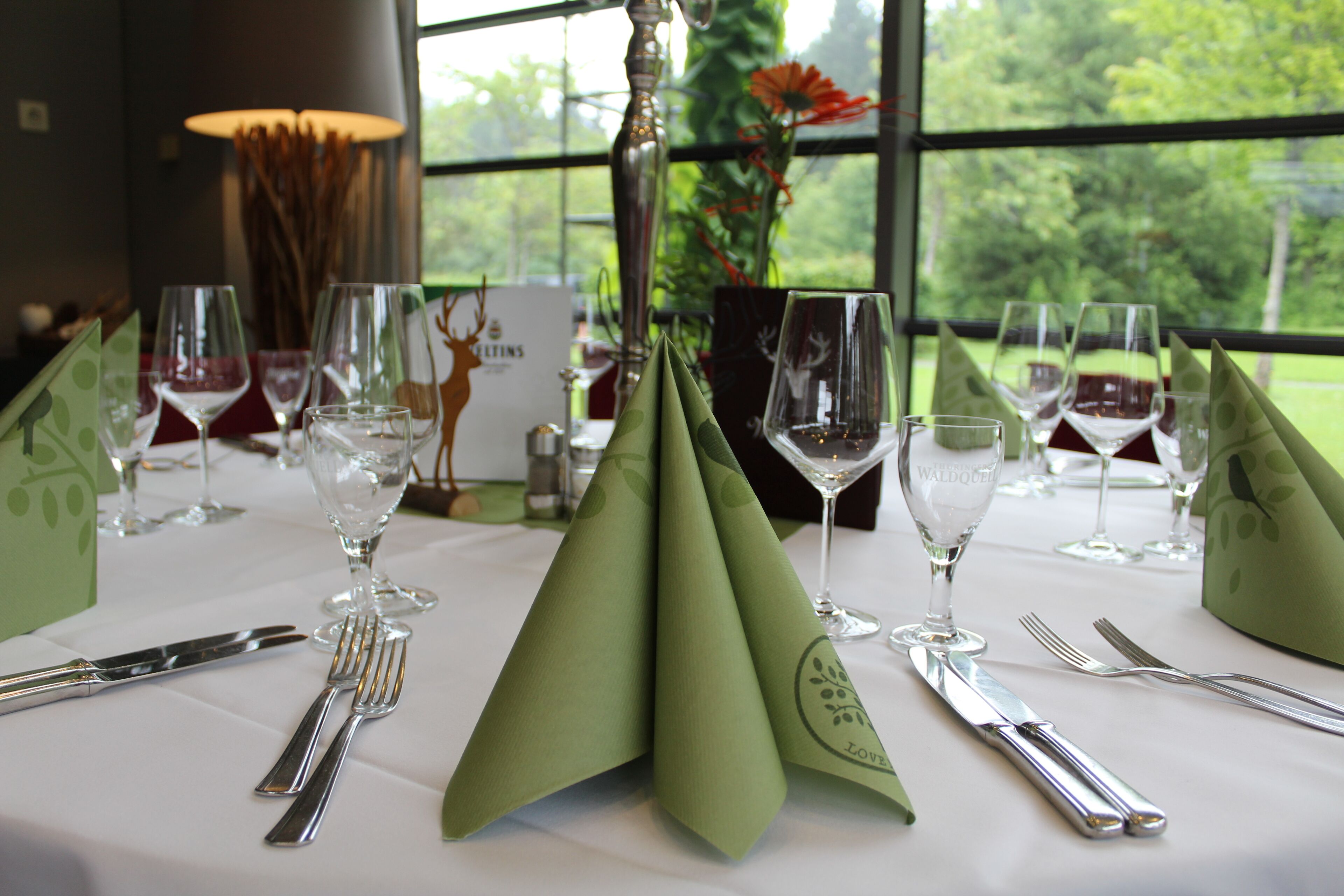 A table set for a meal with a green napkin, silverware, and wine glasses, with a view of trees through a window.
