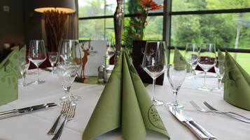 A table set for a meal with a green napkin, silverware, and wine glasses, with a view of trees through a window.