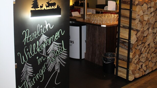 A reception area with a wooden sign, a stack of firewood, and hanging pendant lights. - Hotel Rennsteig (Masserberg)