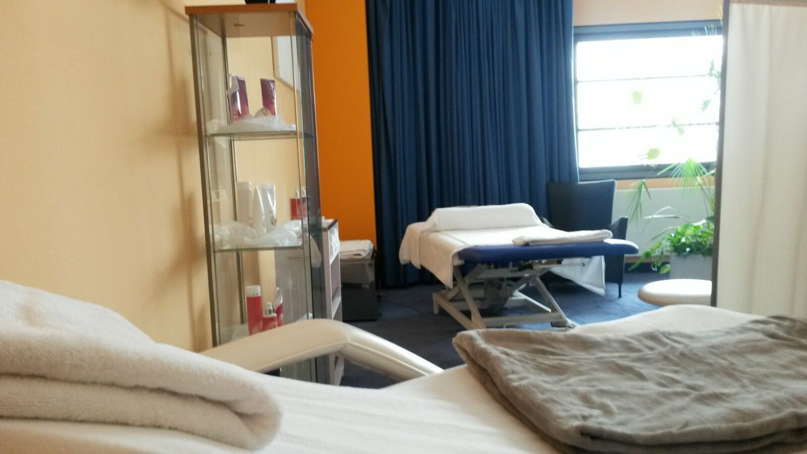 A hotel room with a bed, a massage table, a glass cabinet with toiletries, and a window with curtains.