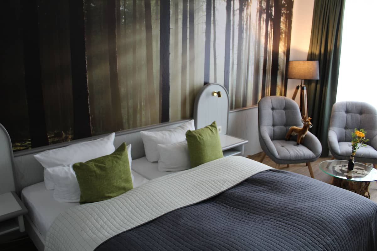 A hotel room with a bed, two armchairs, a small table with a vase, and a large window with curtains.