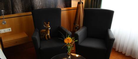 Habitación de hotel con dos sillones negros, una mesita de madera con un florero y una lámpara, además de una estatua de ciervo.