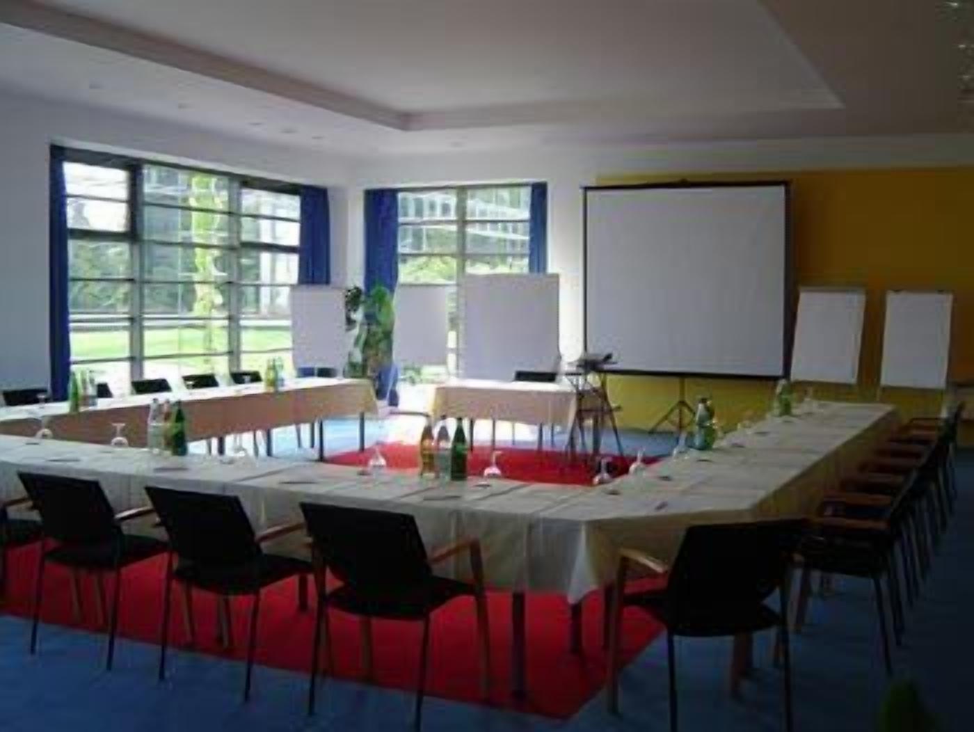 A conference room with a long table, chairs, a projector screen, and windows with blue curtains.