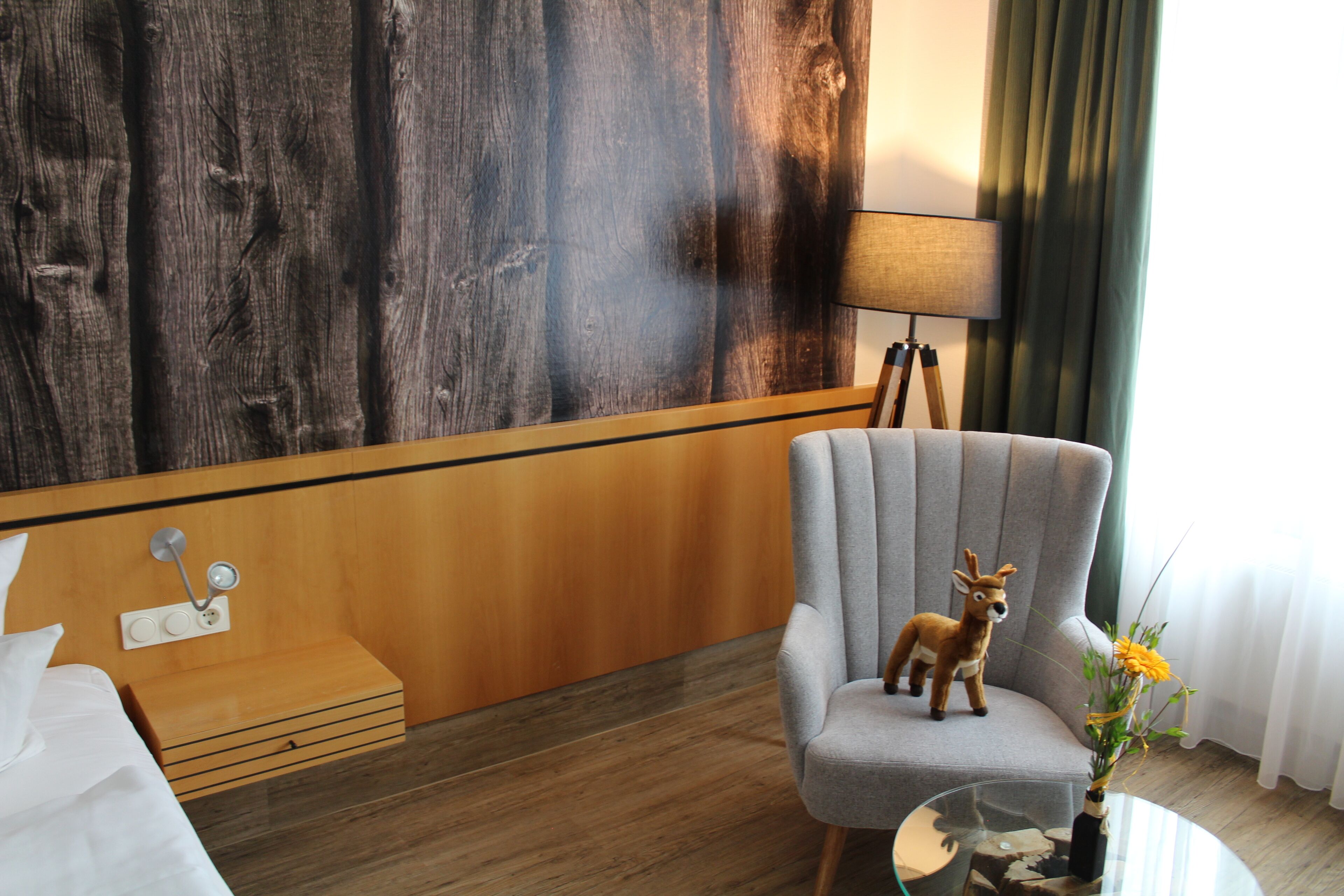 A hotel room with a wooden accent wall, a grey armchair, a small table with a vase of flowers, a lamp, and a bed with white linens.
