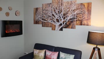 Un salon avec un canapé noir, des coussins colorés, un tableau représentant un arbre accroché au mur, un téléviseur à écran plat et une lampe sur pied.