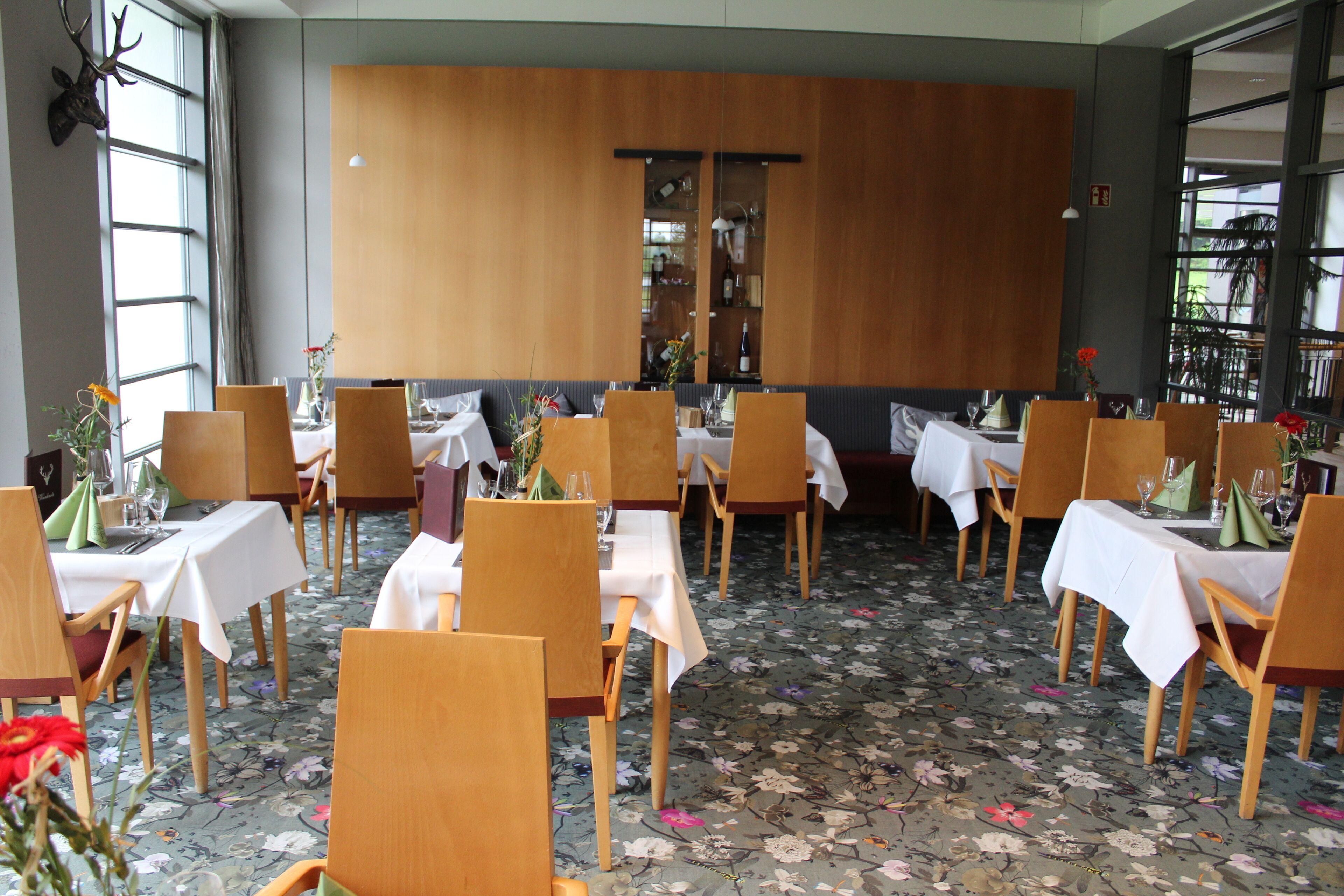 Um restaurante com cadeiras e mesas de madeira, um tapete floral e um grande armário de madeira ao fundo.