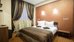 Quarto de hotel com uma cama grande, uma mesa de cabeceira, um painel de madeira na cabeceira, uma janela com cortinas e um termostato na parede.
