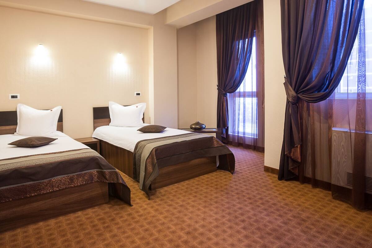 A hotel room with two beds, a large window with curtains, and a carpeted floor.