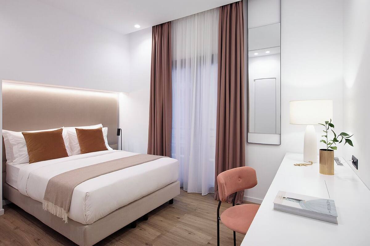 A modern hotel room with a bed, a desk, a chair, and a large window with curtains.