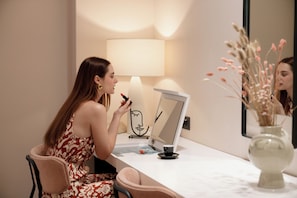 Une femme est assise devant une coiffeuse, devant un miroir, avec une lampe et un vase contenant des fleurs séchées.