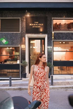 Une femme, vêtue d’une robe à motifs, se tient devant un bâtiment sur lequel on peut lire l’inscription : « THE MARBLOS ATHENS ».