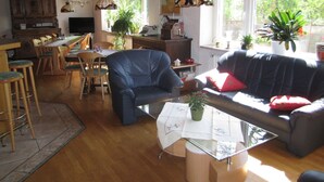 Een ruime woonkamer met een bank, stoelen, een eettafel en een keukenhoek.