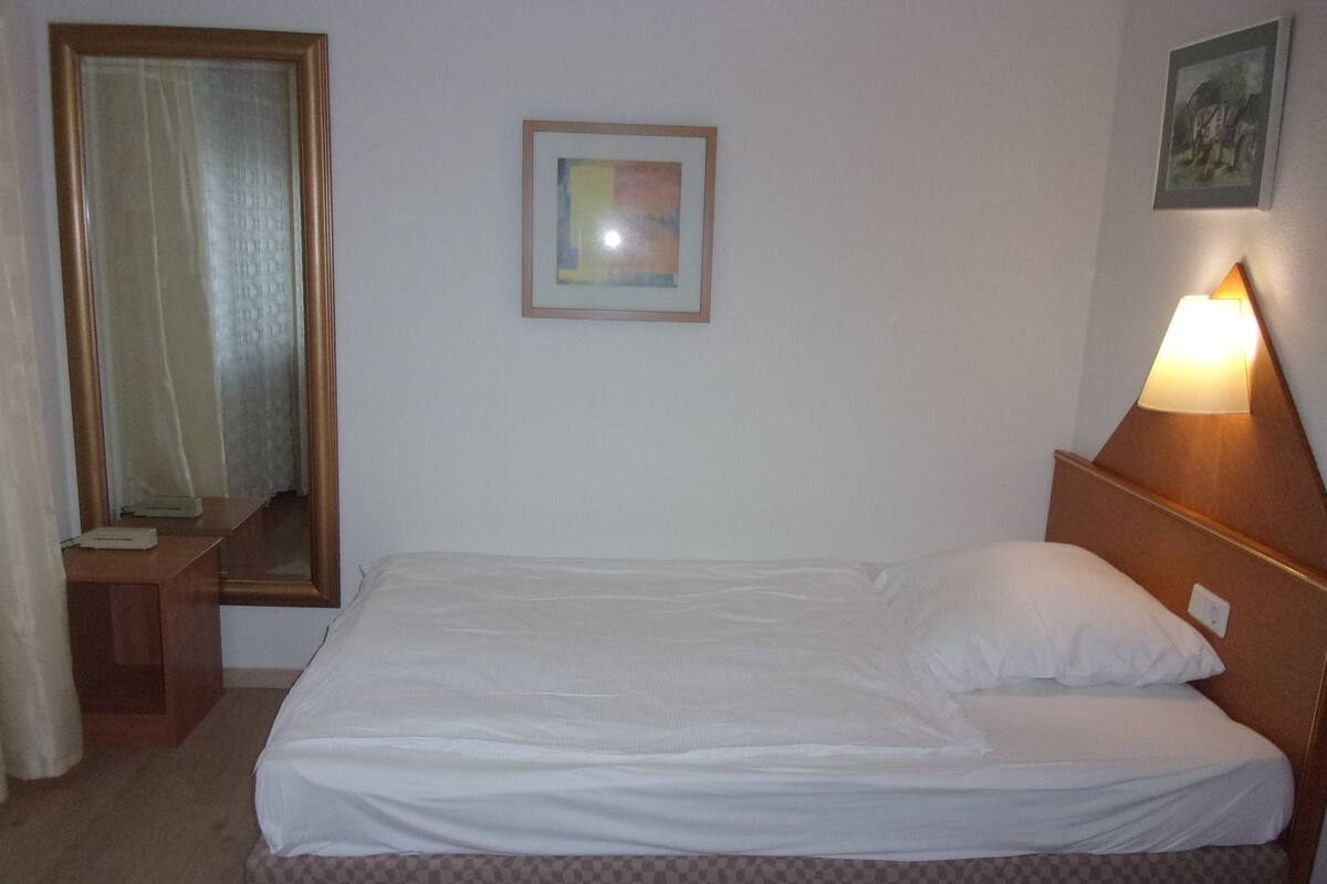 Ein Hotelzimmer mit einem Einzelbett, einer Nachttischlampe, einem Spiegel und einem gerahmten Bild an der Wand.