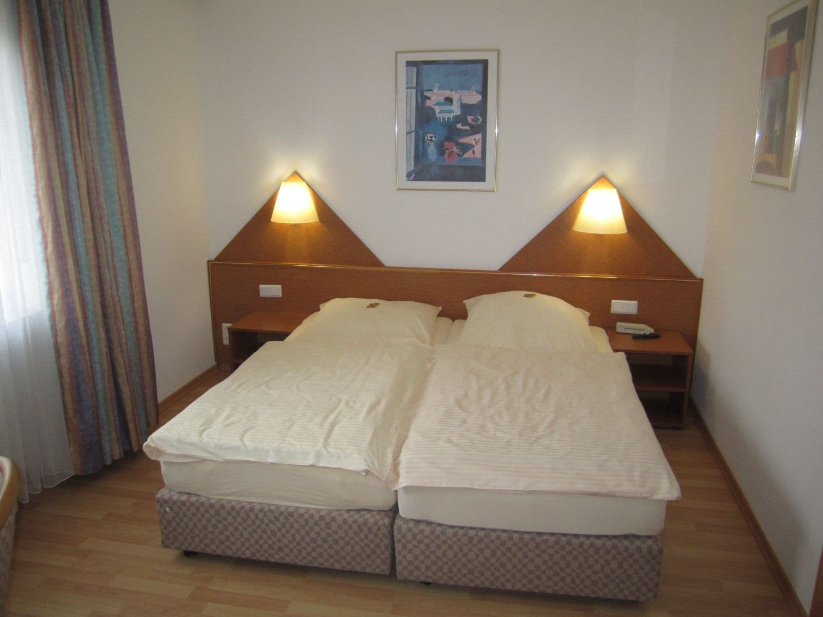 Cama doble con dos mesitas de noche, cada una con una lámpara, en una habitación con suelo de madera y un cuadro enmarcado en la pared.