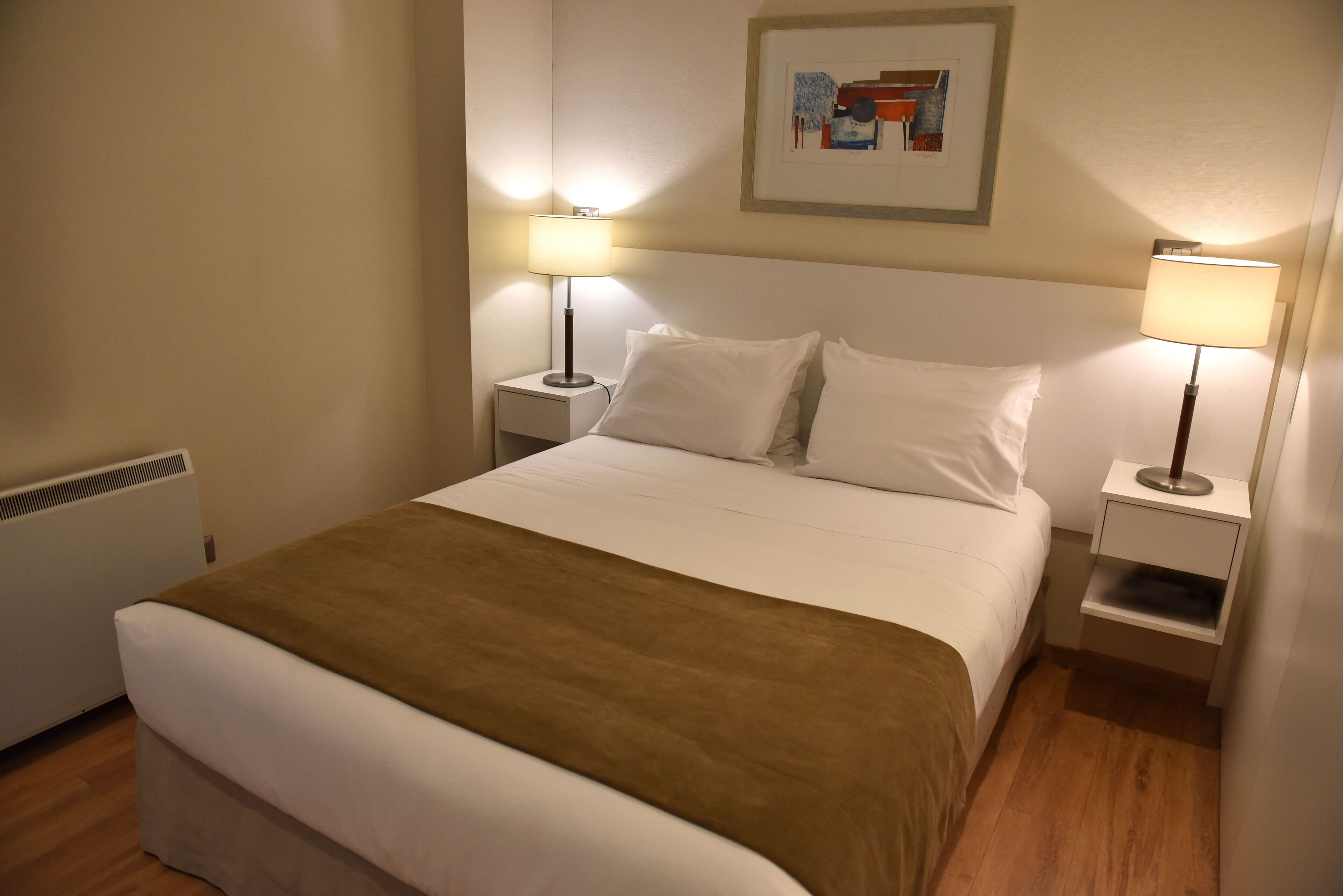 Una cama bien tendida con sábanas blancas y un plaid marrón, flanqueada por dos mesitas de noche con lámparas, en una habitación con radiador y un cuadro en la pared.