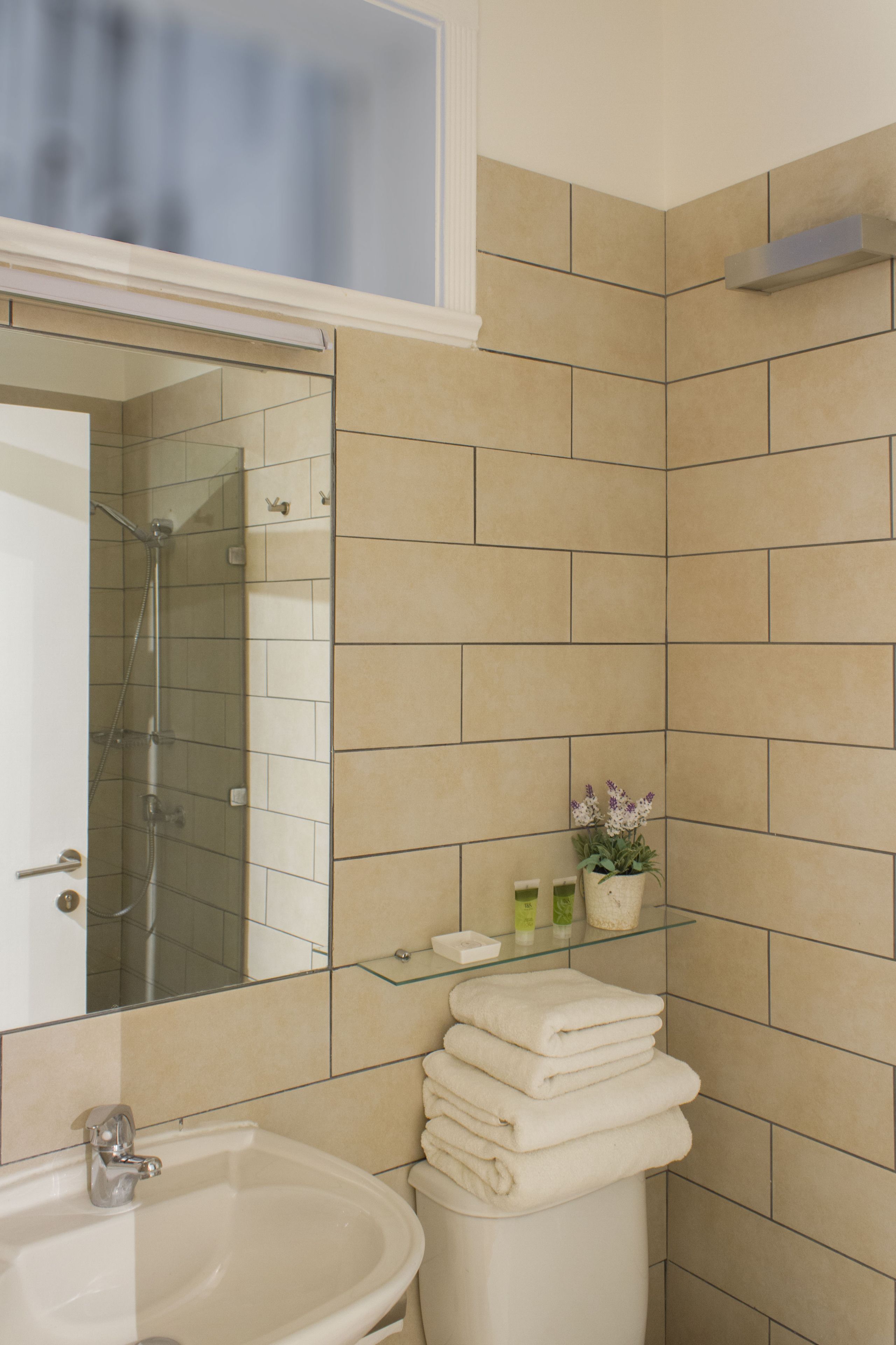 A bathroom with beige tiles, a glass shower enclosure, a sink, and a stack of towels.