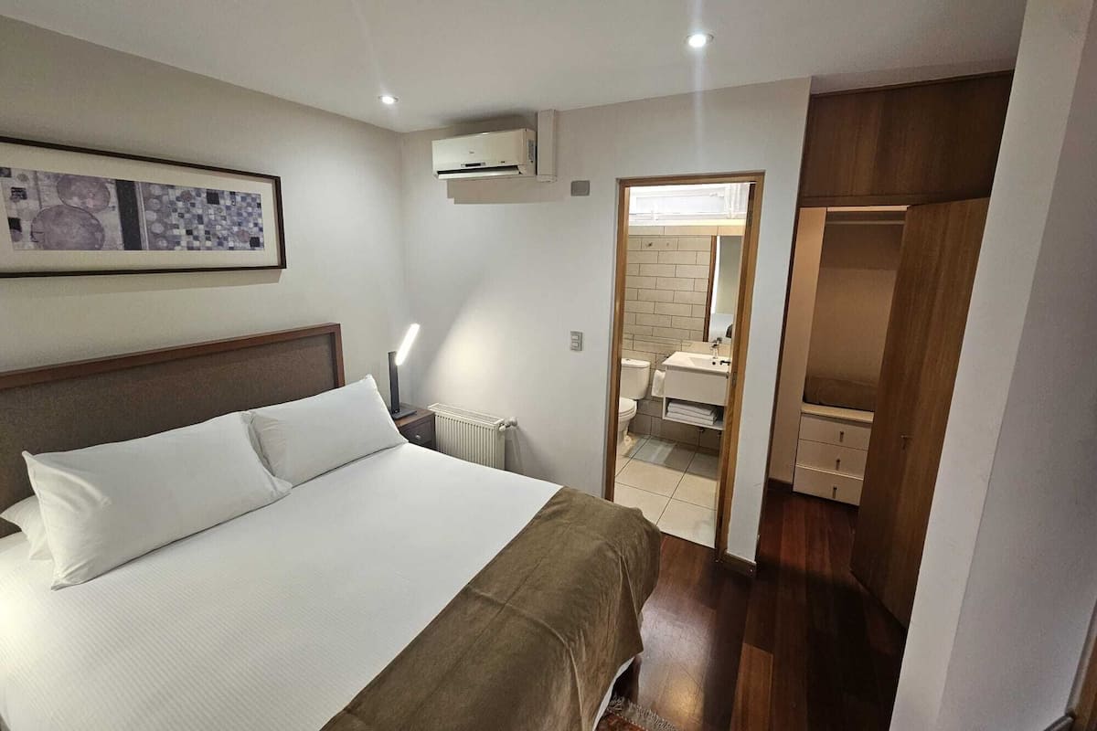 Habitación de hotel con cama, mesita de noche, radiador, unidad de aire acondicionado, baño con inodoro y lavabo, y armario de madera.