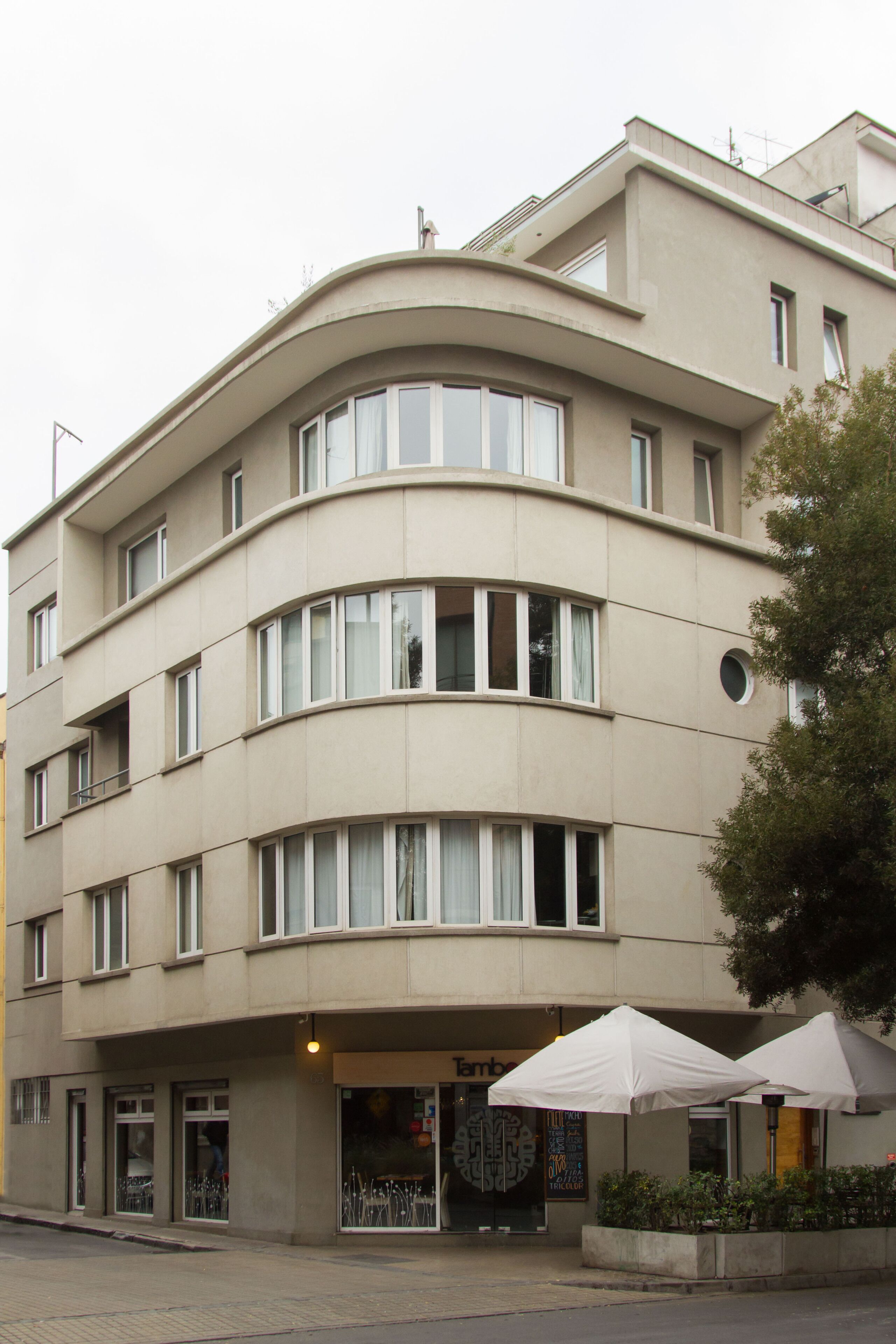Un edificio de varios pisos con fachada curva, ventanas amplias y mesas al aire libre con sombrillas.