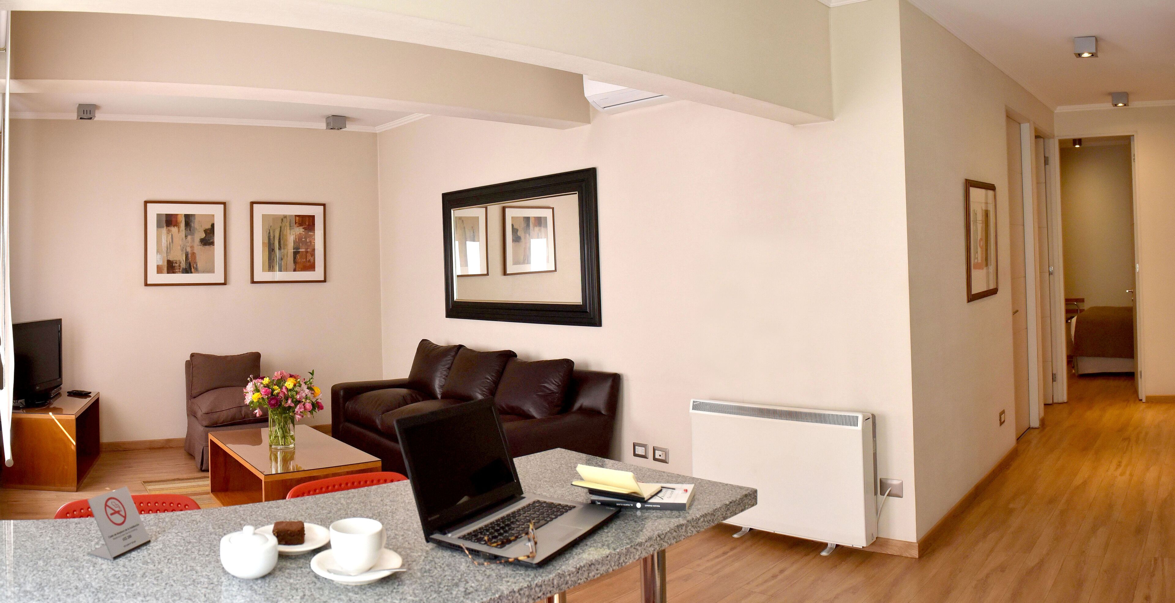 A modern hotel room with a brown leather sofa, a small table with a laptop, a coffee cup, and a vase of flowers. There are framed pictures on the wall and a radiator on the wall.
