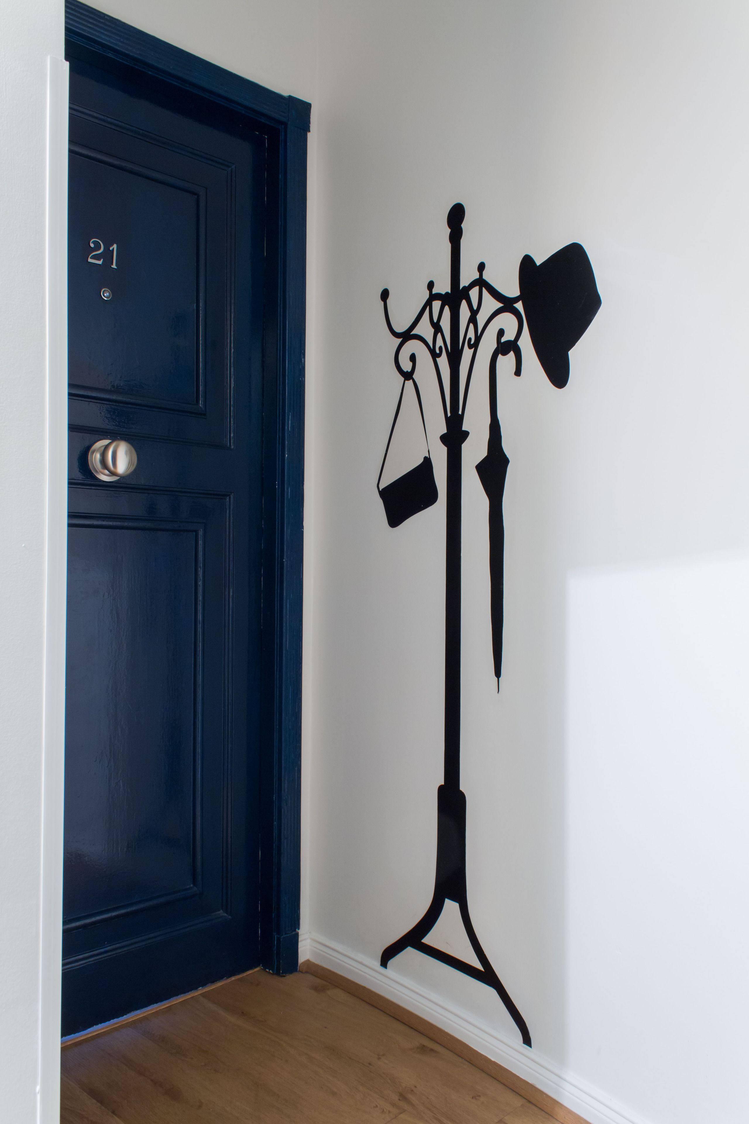 Una puerta azul oscuro con el número 21, un perchero negro con un sombrero y una bolsa, y una pared blanca.