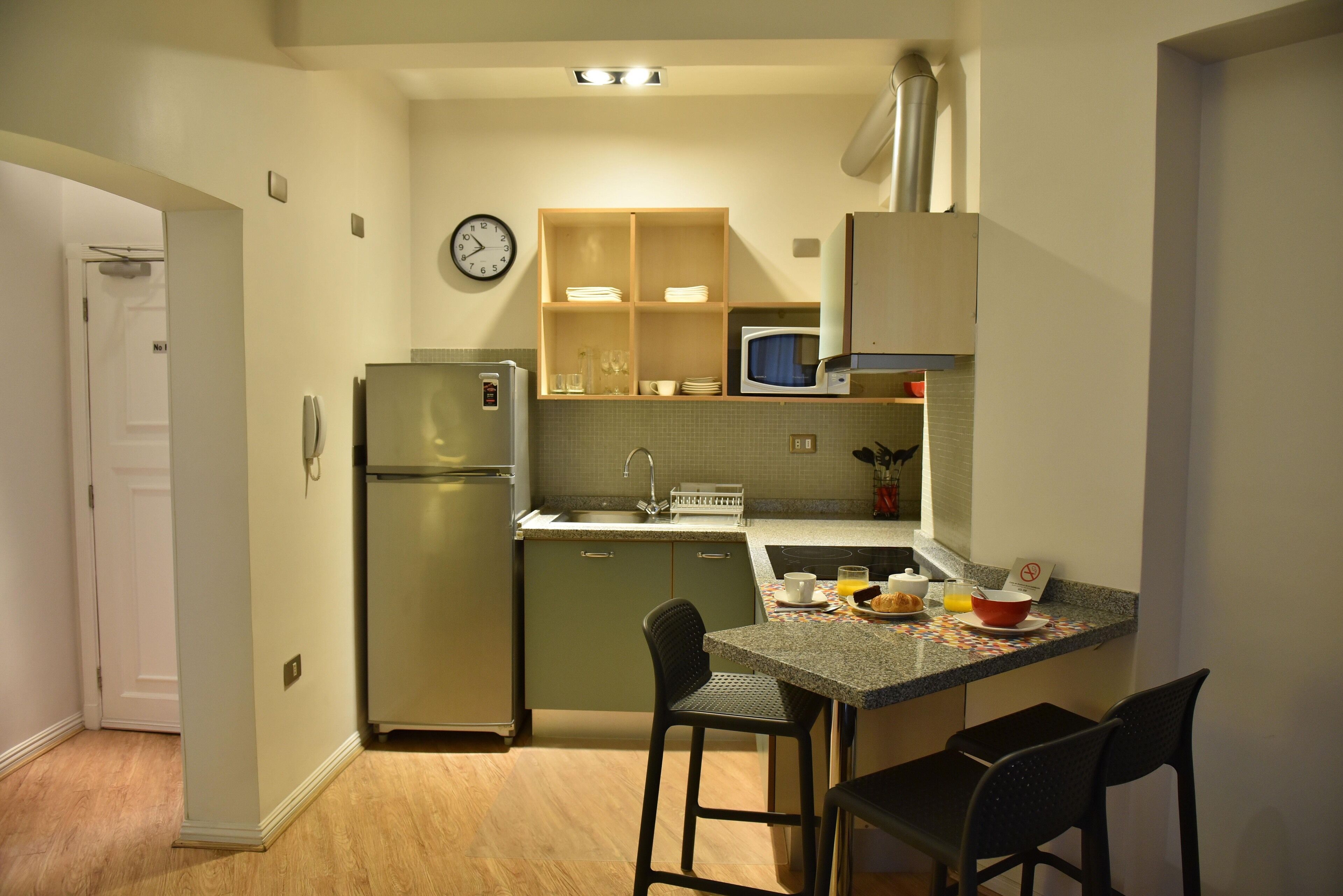 Cocina compacta con refrigerador, microondas y lavamanos. Hay dos taburetes de bar y una mesa pequeña con alimentos para el desayuno.