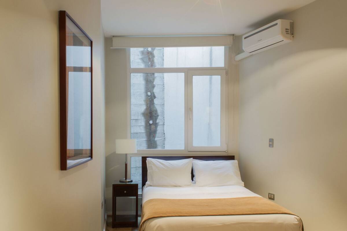 Un dormitorio pequeño y ordenado con una cama individual, una mesita de noche, una lámpara y una ventana que da a un árbol.