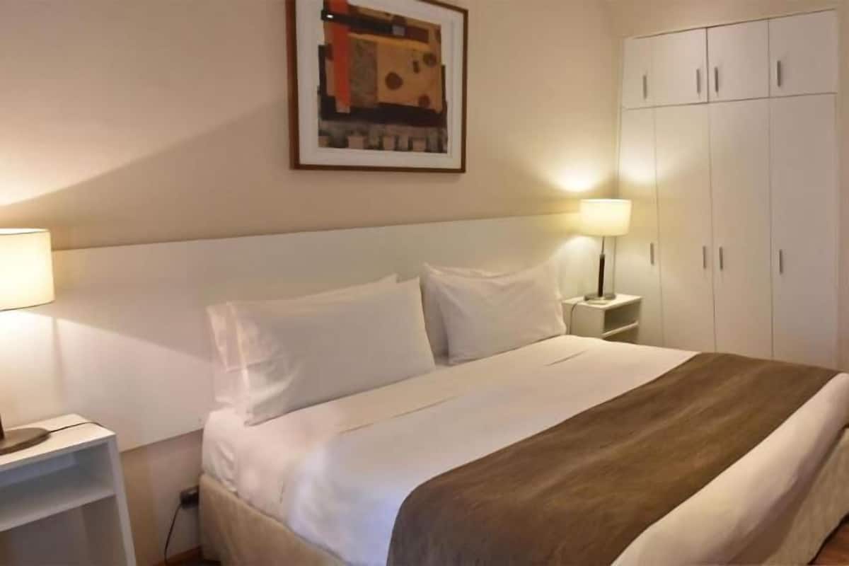 Una cama bien hecha con sábanas blancas y una colcha marrón, una mesita de noche con lámpara, un armario con varias puertas y un cuadro en la pared.