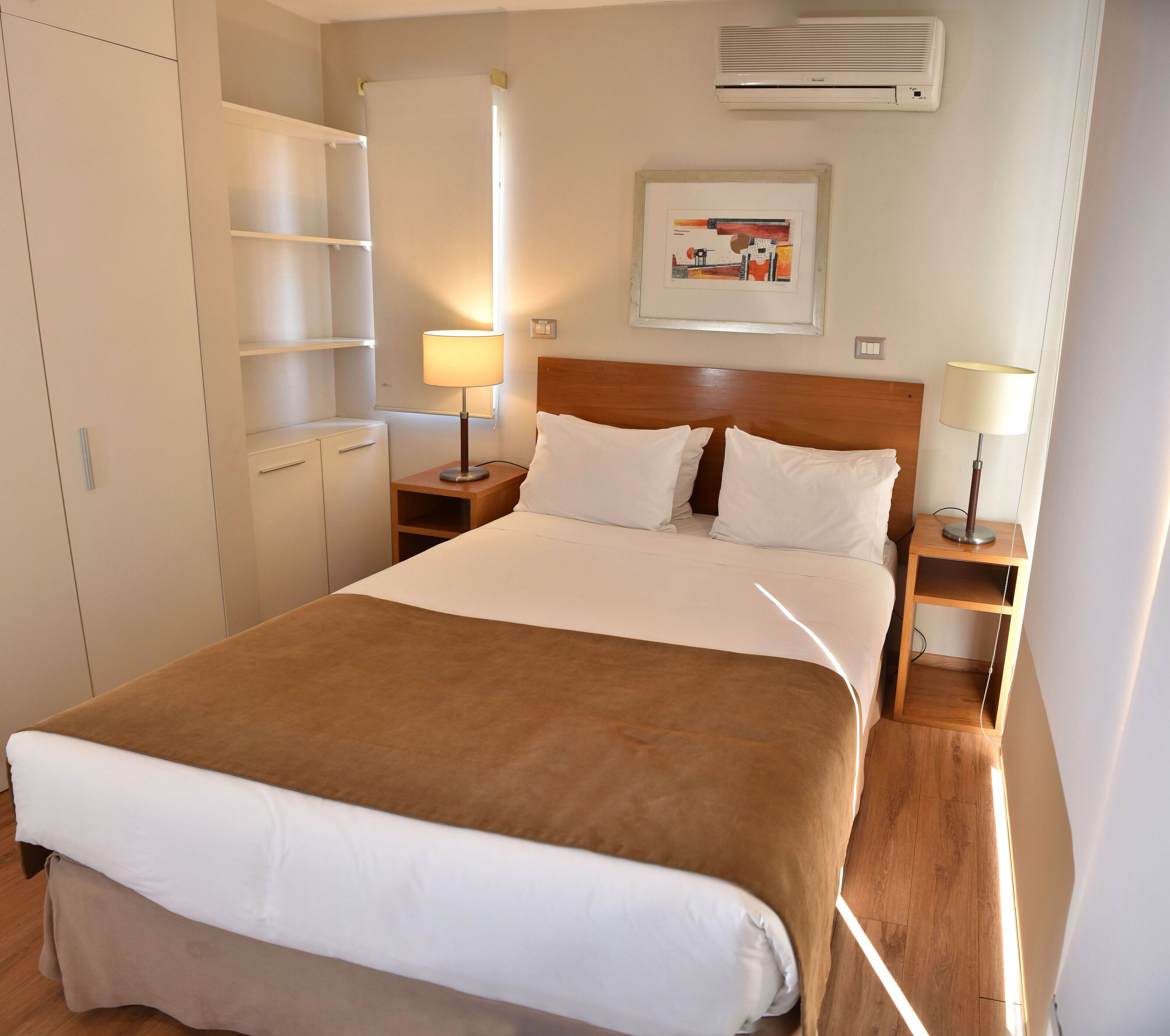 A hotel room with a large bed, two bedside tables with lamps, a wooden headboard, a wardrobe, and an air conditioning unit on the wall.