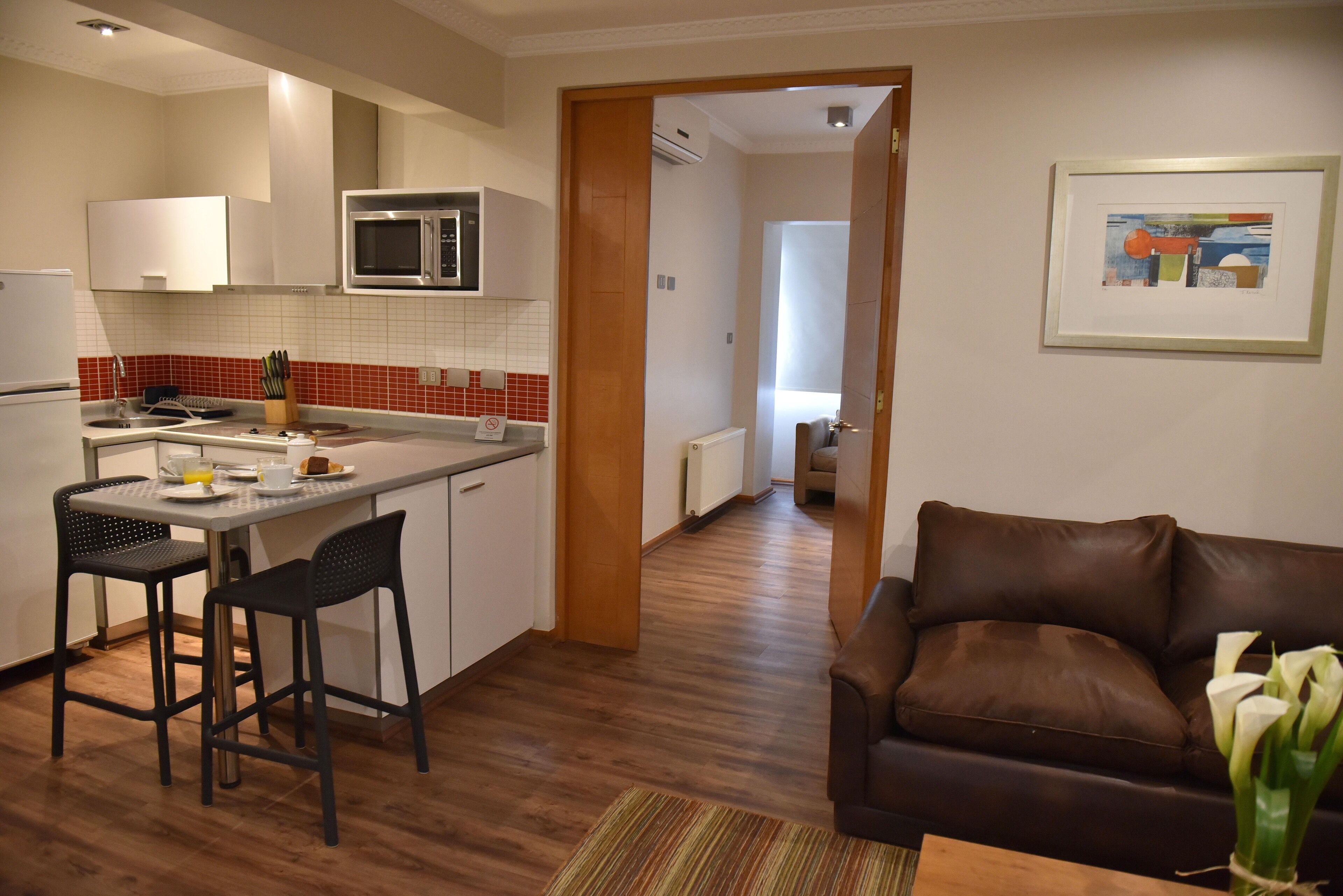 A modern kitchen with a microwave, a dining area with two chairs, and a brown leather sofa in a living room.