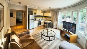 A modern living room with a sofa, a coffee table, and a TV. There is a kitchen with a microwave and a refrigerator. The room has large windows with curtains. - Lastarria Aparts (Santiago)