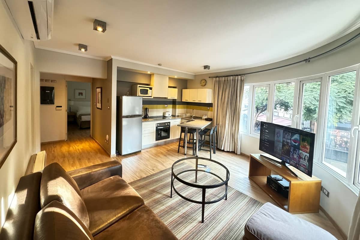 Una sala moderna con sofá, una mesita de centro y televisor. Hay una cocina con microondas y refrigerador. La habitación tiene ventanas amplias con cortinas.